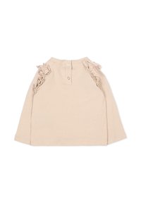 Haut long beige clair avec des accents de volants aux épaules et une fermeture à boutons dans le dos. Tissu doux avec une texture lisse.