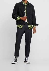 Schwarze Jeansjacke, grün und schwarz karierte Hemd darunter, dunkle strukturierte Hosen und schwarz-weiße Sneakers im Low-Top-Stil.
