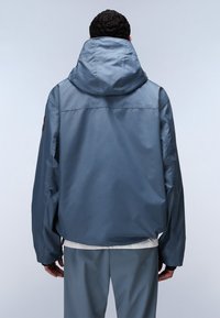 Light blue jacket with a large hood, loose fit, elastic cuffs, and a visible logo patch on the left sleeve. Smooth, weather-resistant material.