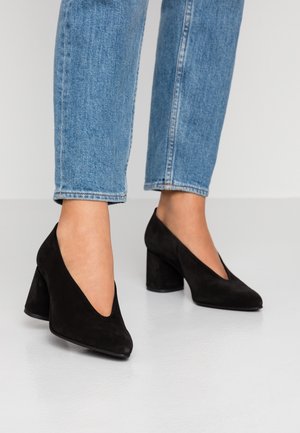 Femme portant un jean bleu et des chaussures noires en daim à talon carré bas, debout sur un sol blanc.