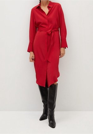 Kobieta ubrana w czerwoną midi sukienkę z długim rękawem i paskiem, w połączeniu z czarnymi długimi kozakami, stojąca na tle jednolitej białej ściany.