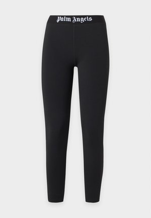 Frau steht in schwarzem Palm Angels Sport-BH und Leggings mit weißen Sneakers vor einem schlichten hellen Hintergrund.