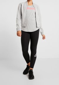 Grå zip-up bomberjakke over en grå t-shirt med rød "adidas" tekst, parret med sorte leggings med en hvid logo og sorte sneakers.