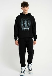 Czarny sweter z grafiką przedstawiającą dwie postacie oraz napisem "EPIPHANY", w zestawieniu z czarnymi spodniami i białymi trampkami z niebieskimi akcentami.