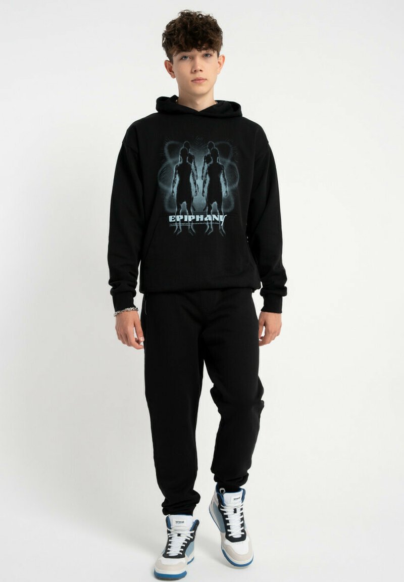 Czarny sweter z grafiką przedstawiającą dwie postacie oraz napisem "EPIPHANY", w zestawieniu z czarnymi spodniami i białymi trampkami z niebieskimi akcentami.
