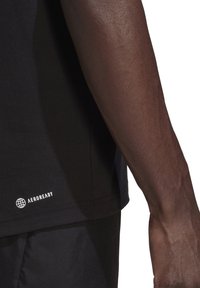 Detailní záběr na tmavě hnědou ruku vedle lemu černého sportovního trička s malým bílým logem Aeroready.