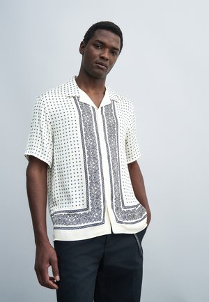 Homme portant une chemise blanche à manches courtes avec des motifs paisley noirs et de petits pois, associée à un pantalon noir sur un fond gris uni.