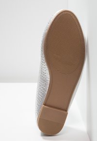 Semelle d'une chaussure plate en caoutchouc beige texturé avec un petit talon, dessus visible en tissu recouvert de paillettes argentées.