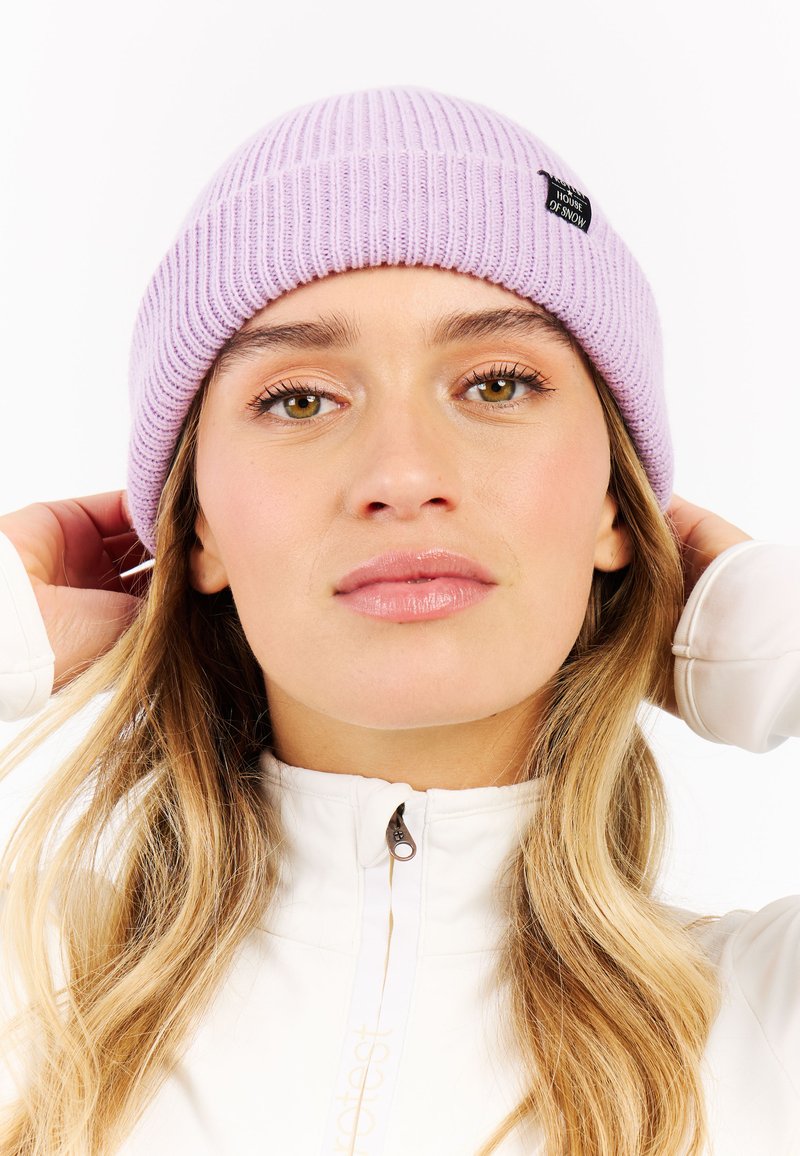 Lavender ribgebreide beanie met een zwart label met daarop "House of Snow." Zachte textuur, snug fit en ronde vorm, gedragen met een witte zip-up jas.