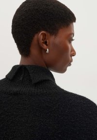 Schwarzer strukturierter Pullover mit hohem Kragen und Rüschendetail auf der Rückseite. Das Model trägt silberne Creolen. Der Hintergrund ist neutral.