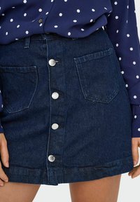 Tumedat pesu denim seelik, ees nupuga kinni klammerdamisega, kahe lisakotiga ja puhta servaga. Kombineeritud tumesinise bluusiga, millel on valged täpid.