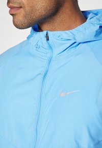 Niebieska lekka kurtka z zamkiem z przodu, wysokim kołnierzem i odblaskowym srebrnym logo Nike na piersi. Gładka tekstura, minimalistyczny design.