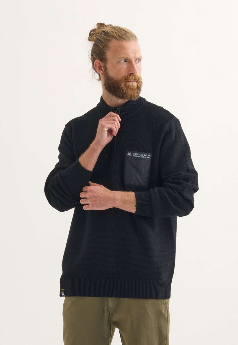 Schwarzer Strickpullover mit hohem Kragen, ausgestattet mit einem Reißverschluss und einer strukturierten Oberfläche. Enthält eine schwarze aufgesetzte Tasche auf der Brust.