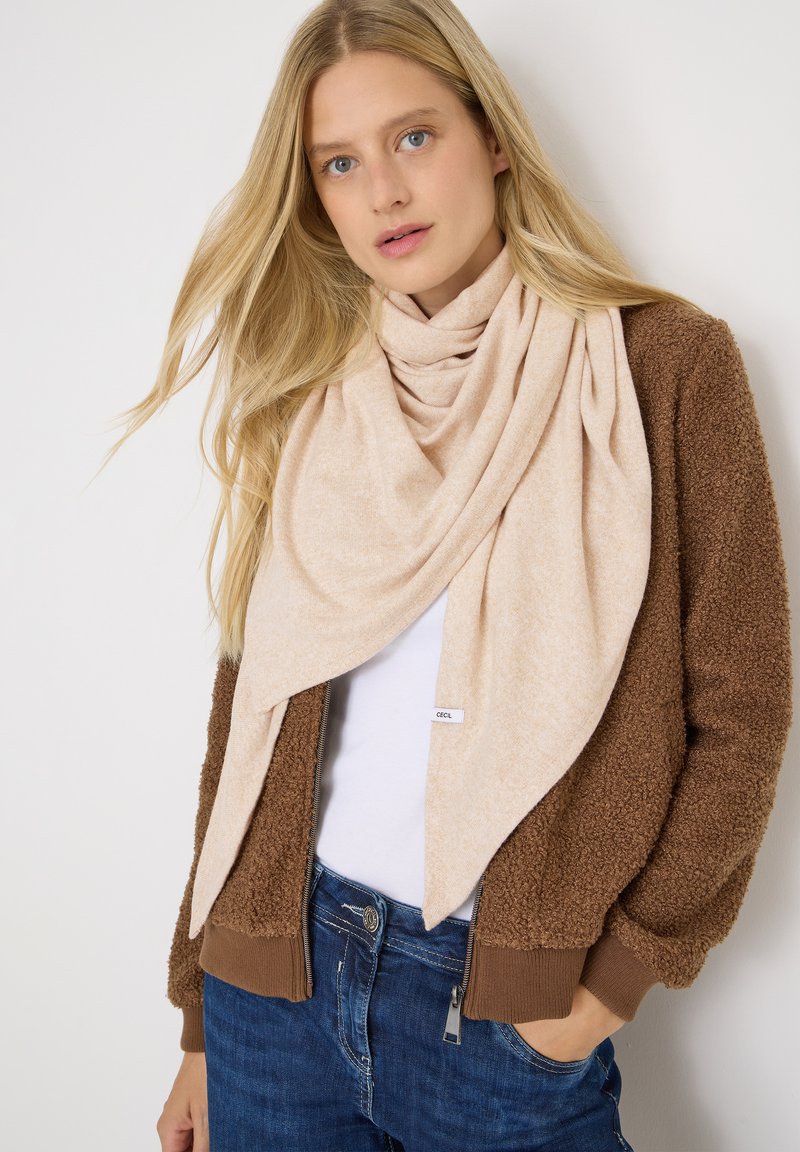 Sciarpa beige con un design morbido e drappeggiato indossata sopra una giacca marrone pelosa. L'outfit è completato da una camicia bianca e jeans blu.