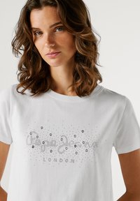 Žena s vlnitými hnědými vlasy, oblečená v bílém tričku se dekorativními stříbrnými cvočky a nápisem "Pepe Jeans London" na přední straně, na jednobarevném pozadí.