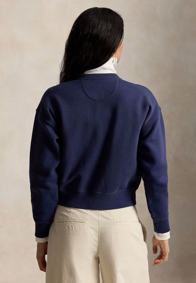 Γυναίκα με μακριά σκούρα μαλλιά, ντυμένη με μπλε σκούνο πουλόβερ πάνω από λευκό ζιβάγκο και μπεζ παντελόνια, που φαίνεται από πίσω σε ουδέτερο φόντο.