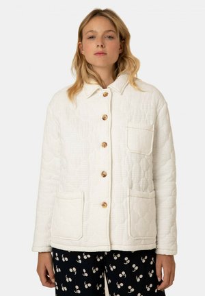 Quilted white jacket with a collared neckline, wooden buttons, and two front pockets; worn over black pants with a cherry pattern.