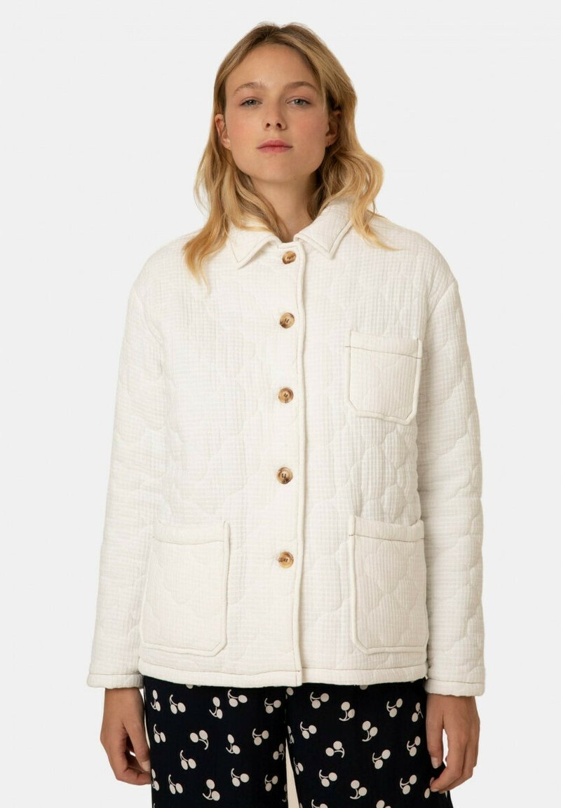 Gequiltete weiße Jacke mit Kragen, Holzknöpfen und zwei Fronttaschen; getragen über schwarzen Hosen mit einem Kirschmuster.