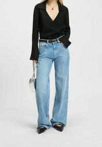 Blouse noire à manches longues de style portefeuille associée à un jean large bleu clair. Comprend une ceinture noire, des chaussures pointues noires et un sac à main argenté.