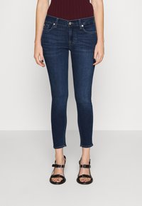 Mujer vistiendo jeans ajustados de color azul oscuro y sandalias negras con hebillas, de pie contra un fondo claro y liso.