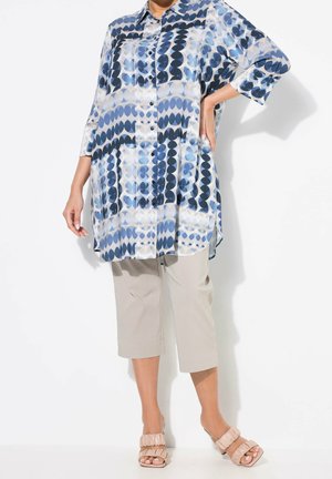 Personne portant une longue chemise bleue et blanche à motifs avec des boutons, un pantalon beige retroussé et des sandales roses clair froncées, debout avec une main sur la hanche.