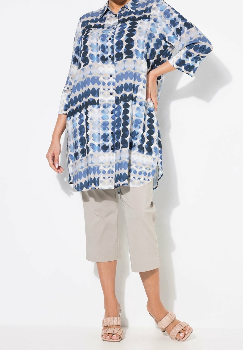 Personne portant une longue chemise bleue et blanche à motifs avec des boutons, un pantalon beige retroussé et des sandales roses clair froncées, debout avec une main sur la hanche.