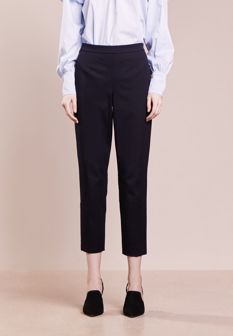 Personne portant un pantalon noir court, un chemisier bleu clair à manches longues et des chaussures noires à talons, debout sur un fond neutre.