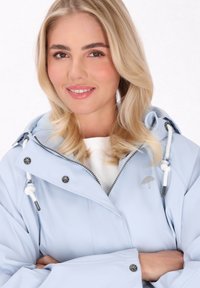 Šviesiai mėlyna atspari vandeniui striukė su kapišonu, su sidabro akcentais, raišteliais ir siuvinėtu skėčio logotipu ant krūtinės.