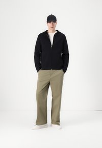Svart zip-up hoodie över en vit t-shirt, ihop med avslappnande olivgröna byxor och vita sneakers, avslutad med en svart keps.
