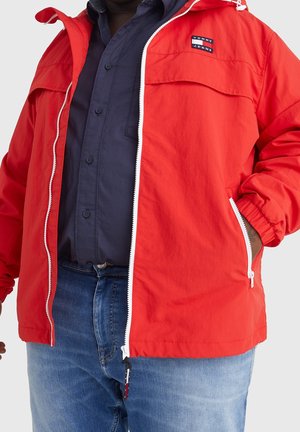 Osoba nosząca czerwoną kurtkę Tommy Jeans na ciemnej koszuli zapinanej na guziki i niebieskich dżinsach, z jedną ręką w kieszeni kurtki.