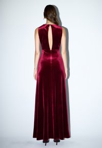 Satinbordeaux kjole med høj hals og åben ryg. Stoffet er glat og strømmer ned til gulvet med en elegant silhuet.