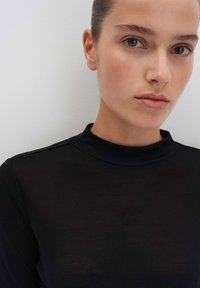 Jonge vrouw met een lichte huid en teruggebonden haar, gekleed in een zwarte hoge nek top tegen een effen lichte achtergrond.