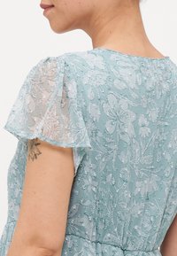 Femme portant une robe florale légère bleu clair avec des manches volantes et un petit tatouage visible sur le haut du bras.