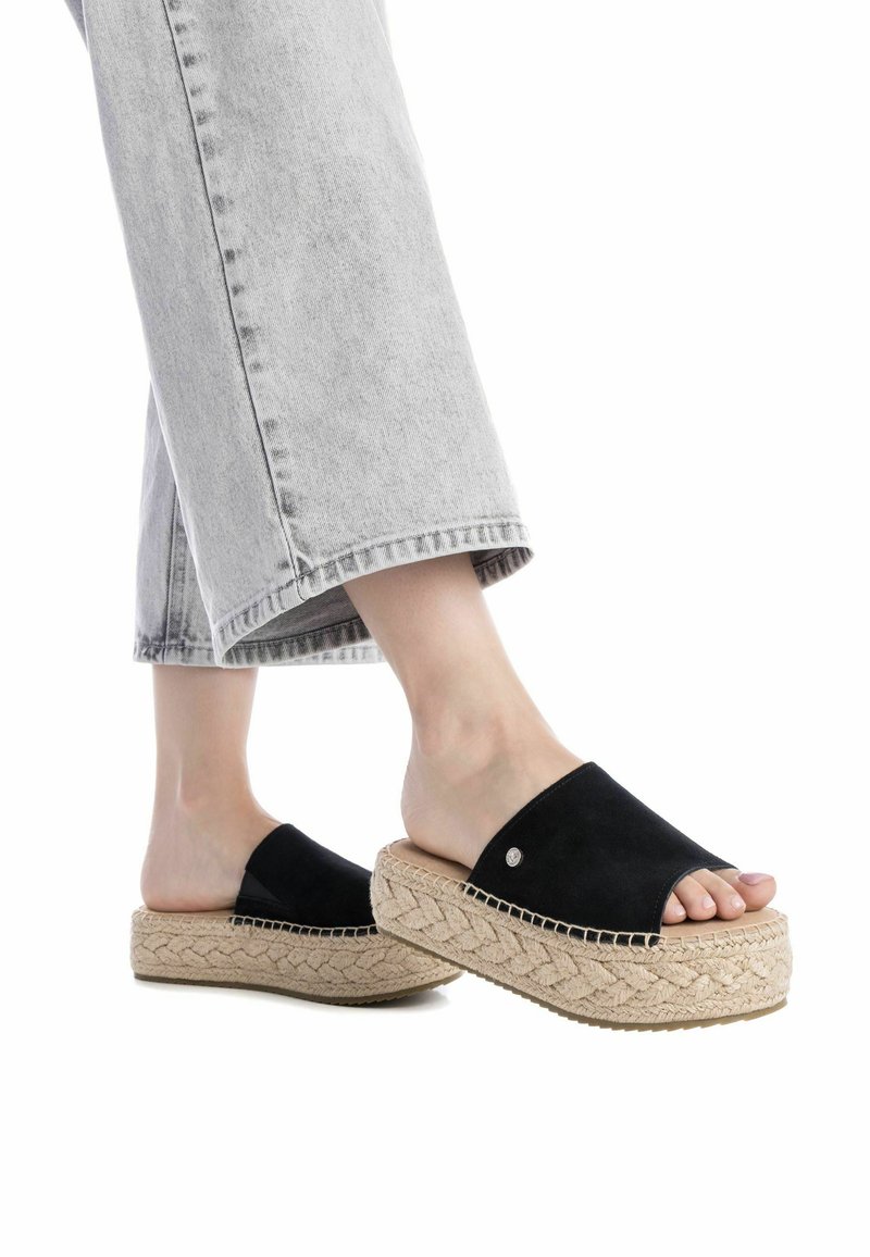 Sandalias de plataforma de ante negro con puntera destapada, que presentan una suela media trenzada similar al yute y un sutil acento metálico, combinadas con pantalones recortados en gris claro.