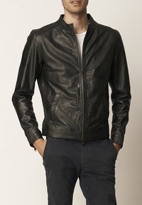Chaqueta de cuero negra con cuello alzado, cierre frontal con cremallera y dos bolsillos laterales. Textura suave con un ligero brillo y corte ajustado.