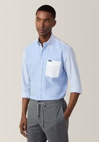 Camisa de botón azul claro con mangas a rayas, presenta un bolsillo en el pecho blanco, combinada con pantalones grises con cordón. Ajuste y diseño casual.