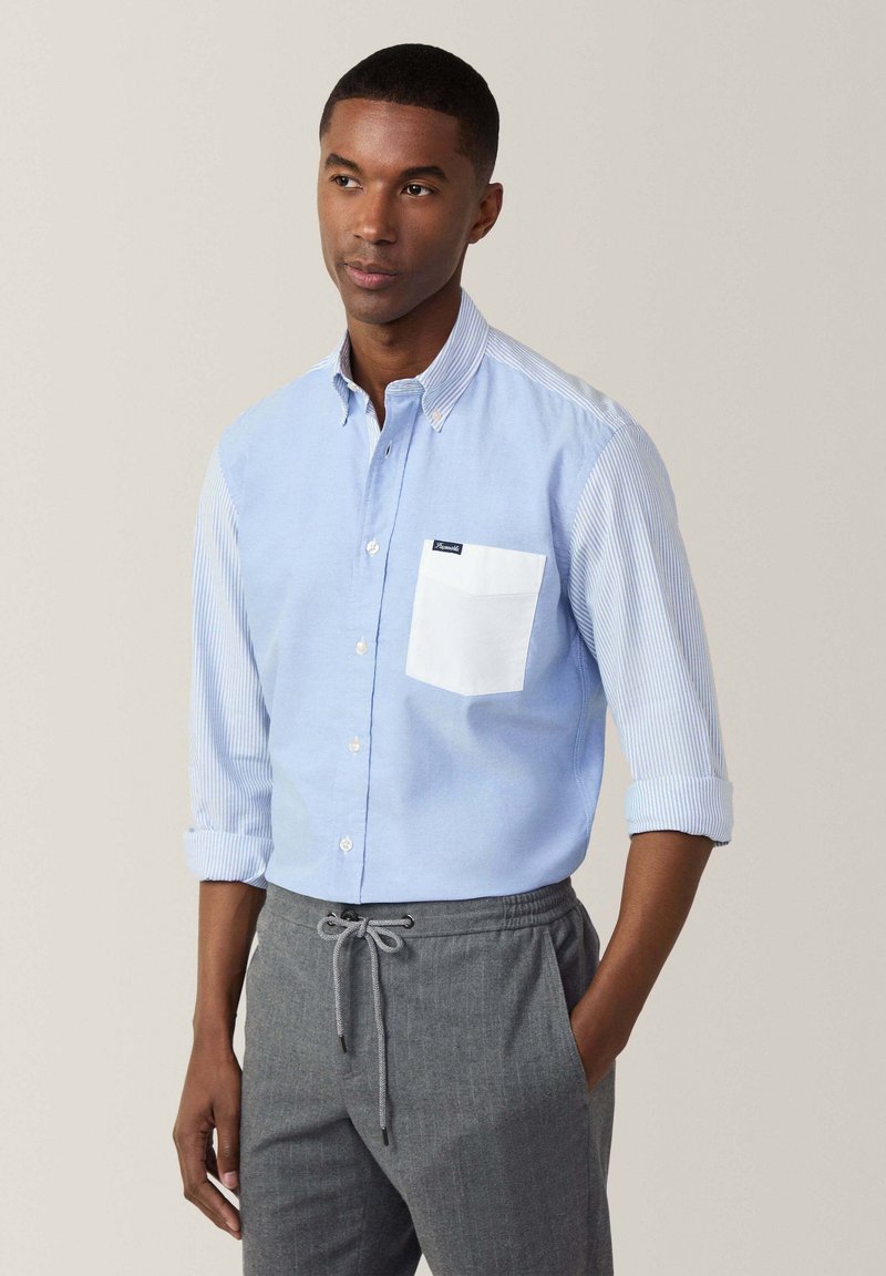 Camisa de botón azul claro con mangas a rayas, presenta un bolsillo en el pecho blanco, combinada con pantalones grises con cordón. Ajuste y diseño casual.