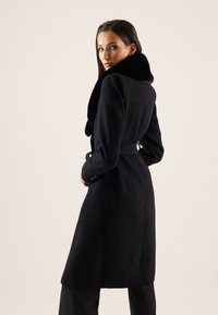 Femme aux longs cheveux bruns portant un manteau noir ceinturé avec un grand col en fourrure, regardant par-dessus son épaule sur un fond uni.