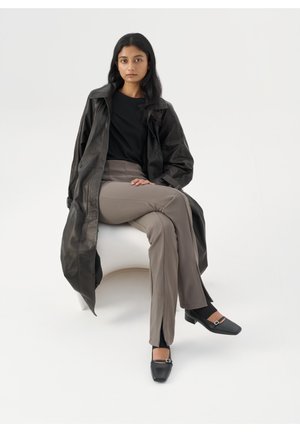 Manteau en cuir noir, haut noir, pantalon gris ajusté et chaussures noires à bout carré avec boucles, sur un fond blanc minimaliste.
