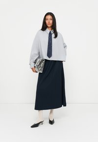Helles graues, übergroßes Hemd mit einer dunkelblauen Krawatte, kombiniert mit einem plissierten, marineblauen Rock und schwarzen spitzen High Heels. Die Accessoires umfassen eine Tasche mit Schlangenprint.