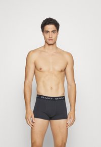 Boxeri negri din material moale și elastic, cu un brâu cu logo alb în contrast. Modelul stă cu o expresie neutră pe un fundal simplu.