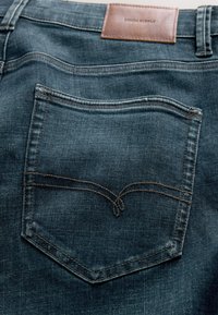 Jean en denim bleu foncé avec poche arrière cousue et étiquette en cuir marron indiquant "Denim Supply" à la taille.