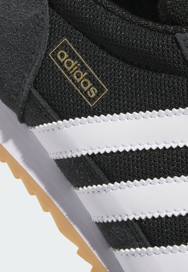 Detail eines schwarzen Adidas-Sneakers mit strukturierter Oberfläche, weißen drei Streifen, Gummisohle und goldfarbenem Adidas-Logo an der Seite.