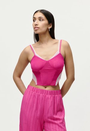 Kvinde med langt mørkt hår iført en pink cropped camisole og højttaljede pink bukser, der kigger til venstre med hænderne bag ryggen.