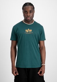Zelené bavlněné tričko s krátkým rukávem, na kterém se ve středu nachází žlutý potisk s logem "Alpha Industries" a kulatým výstřihem.