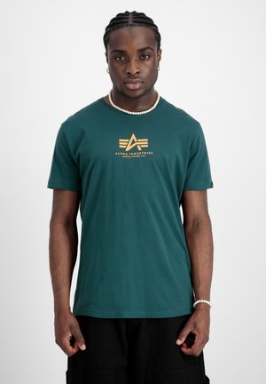 Zöld pamut póló rövid ujjakkal, középen sárga "Alpha Industries" logónyomattal és kerek nyakkivágással.