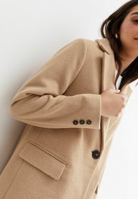 Manteau beige en mélange de laine avec un col cranté, une fermeture à un bouton et des poignets à trois boutons. Comprend une poche latérale et une texture douce.