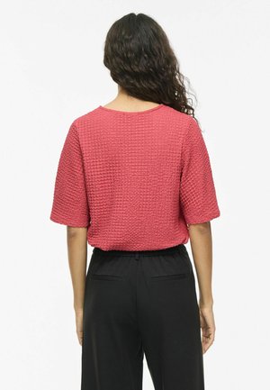 Femme aux longs cheveux ondulés, portant un haut rose texturé à manches courtes et un pantalon noir, vue de dos sur un fond blanc uni.