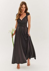 Dlhé, tmavošedé šaty s prekrývacím dizajnom, krátkymi rukávmi a zhrnutým pásom, vyrobené z hladkej látky. Modelka drží biele kala.