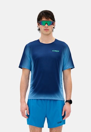 Junger Mann trägt ein blaues Gradient-Sportshirt und Shorts, reflektierende Sportsonnenbrille und eine schwarze Smartwatch, steht vor weißem Hintergrund.